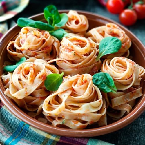 A close-up of beautifully presented pasta nests in a terracotta dish, garnished with fresh basil leaves and cherry tomatoes in the background.