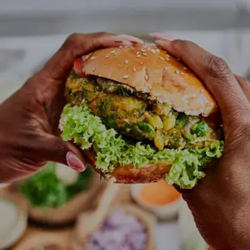 Hands holding a veggie burger with a sesame seed bun, lettuce, and a vegetable patty