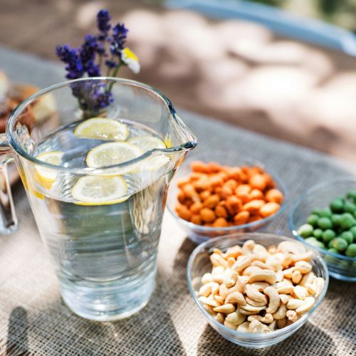 A clear glass pitcher filled with water and lemon slices on a table, along with bowls of assorted nuts and snacks. There is a small vase with flowers in the background.