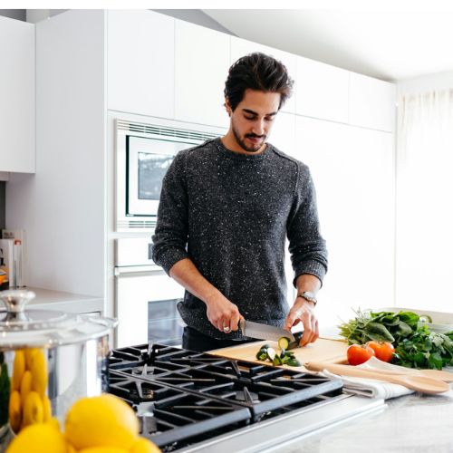 A man in a modern kitchen cutting vegetables on a wooden cutting board. The countertop has fresh produce, utensils, and a large pot on the stove, with orange pots on the other counter.