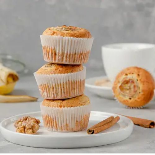 Three banana muffins stacked on a white plate with walnuts and cinnamon sticks on the side; a mug and a banana in the background