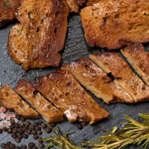 Sliced seitan strips on a dark surface, garnished with pink Himalayan salt, black peppercorns, and sprigs of rosemary