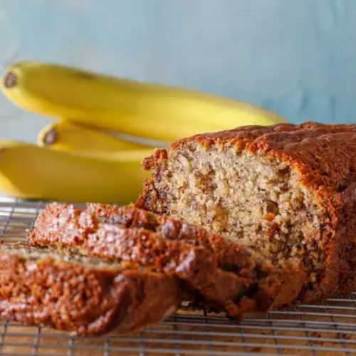 A loaf of freshly baked banana bread cut into slices, placed on a cooling rack with ripe bananas in the background