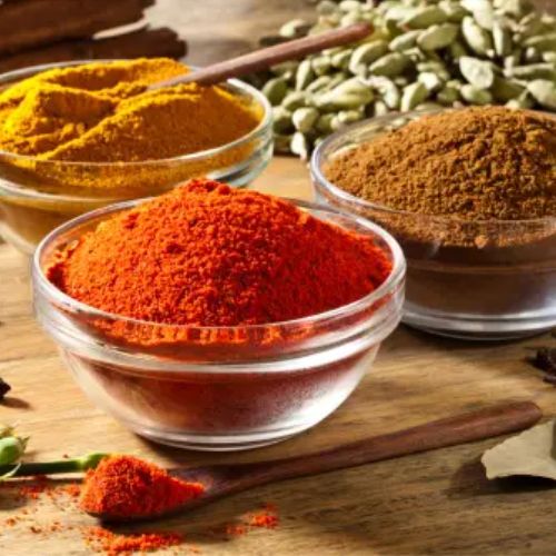 Three glass bowls filled with red, yellow, and brown spices, surrounded by various whole spices and herbs on a wooden table