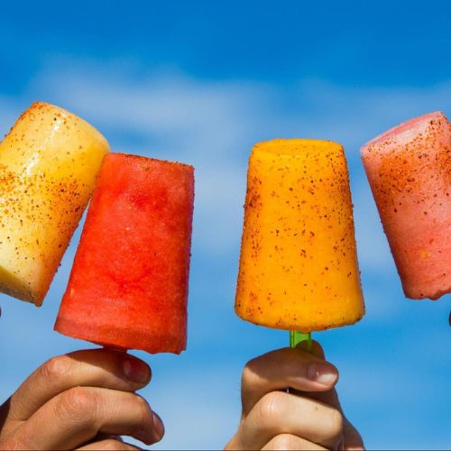 Close-up of hands holding various Mexican popsicles in different flavors and colors with a clear blue sky in the background.