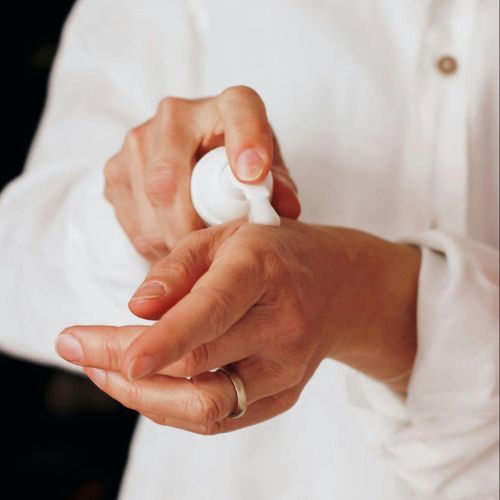 A close-up of a person applying lotion to their hands through a pump dispenser, wearing a white shirt.