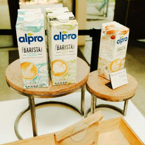 Three cartons of Alpro Barista plant-based milk alternatives (Coconut, Soya, and another flavor) displayed on two wooden round stands in a retail setting with a glass door in the background.
