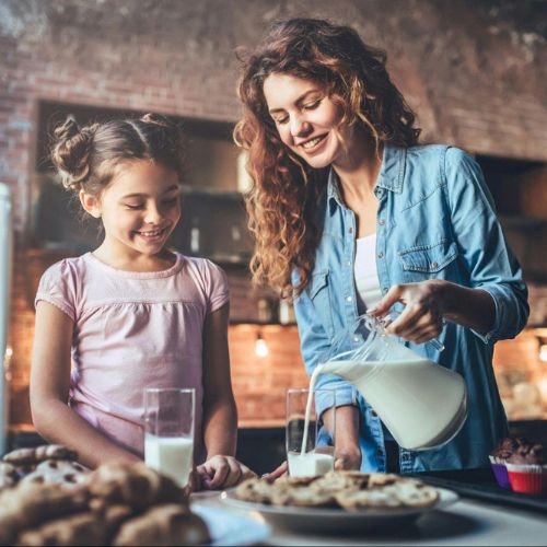 A woman and a young girl smiling and standing in a kitchen, with the woman pouring milk from a pitcher into a glass. Various baked goods such as cookies and muffins are on the counter in front of them.