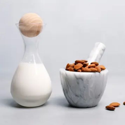 A clean, minimalist setup featuring a glass carafe of milk with a wooden stopper and a marble bowl filled with almonds on a light gray background.