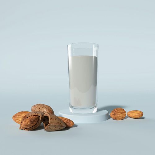 A tall glass of almond milk standing on a small pedestal, surrounded by whole almonds and almond shells, set against a light blue background.