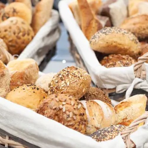 Three wicker baskets lined with white cloth are filled with a variety of freshly baked bread rolls and buns, including seed-covered and plain varieties.