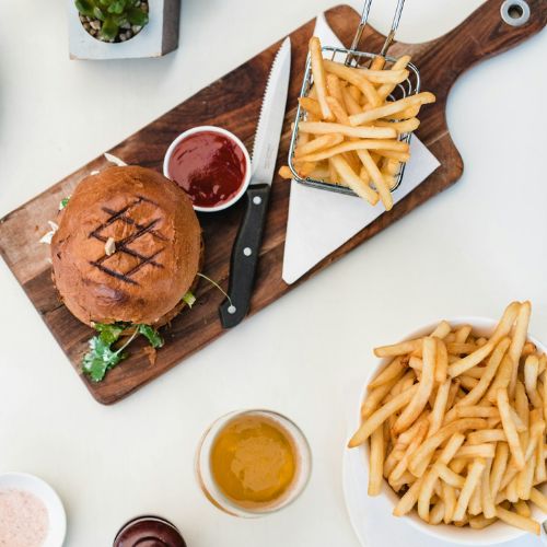 An overhead view of a meal spread featuring a burger on a wooden board with a knife and fork, a small basket of French fries with ketchup on the side, a bowl of calamari, a drink, and a small plant. 