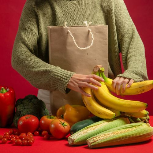 A person unpacking groceries from a brown paper bag, with a variety of fresh produce including bananas, corn, tomatoes, cucumbers, bell peppers, and broccoli displayed on a red surface. The person is wearing a green sweater.