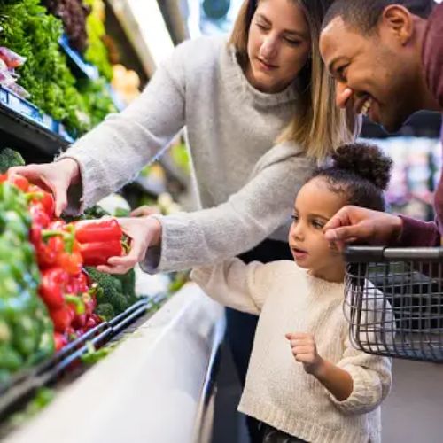 A family picking vegetables in a grocery store. The mother selects a red bell pepper from a display, while the father and child look on and smile. The child points towards the vegetables, engaging in the activity.