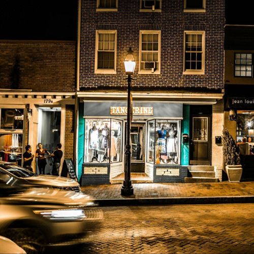 A nighttime street view of a row of shops illuminated by streetlights and store displays. Cars are passing by, and a few people are gathered outside a shop named "TANGERINE." Other visible signs include "Jean Louis" and "CAFÉN."