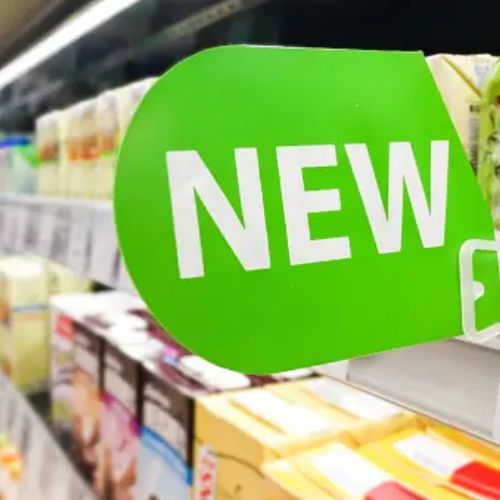 An aisle in a grocery store with shelves stocked with various products. A green sign marked "NEW" is prominently displayed, indicating new items available on the shelf.
