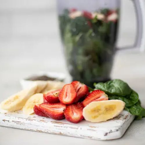 Freshly sliced strawberries and bananas arranged on a cutting board, with a blender in the background containing leafy greens and other smoothie ingredients.