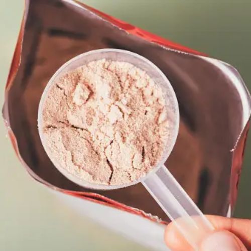 A close-up of a scooper filled with protein powder hovering above an open bag of the same protein powder.