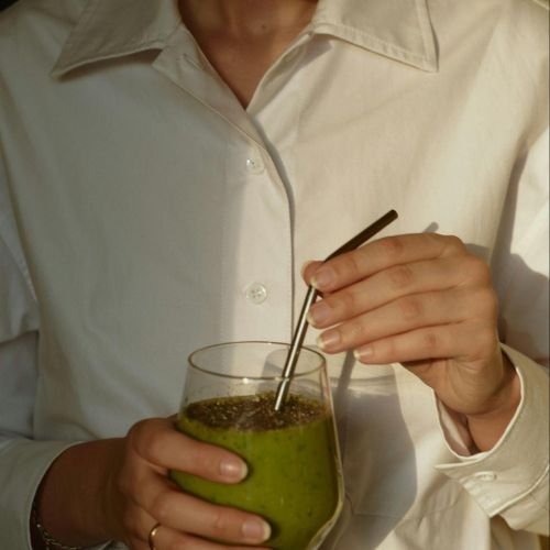 A person in a white shirt stirring a green smoothie in a glass with a reusable metal straw. The smoothie is topped with chia seeds.