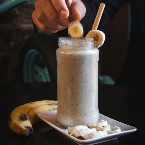 A person adding banana slices to a banana smoothie in a mason jar, with a whole banana and some banana slices on a plate beside it.