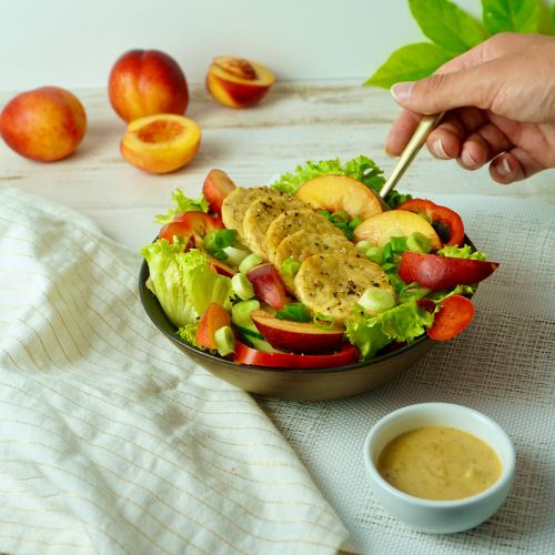 A close-up of a fresh salad in a bowl, topped with sliced grilled chicken and peaches, accompanied by a small dish of dressing. There are additional whole peaches in the background.