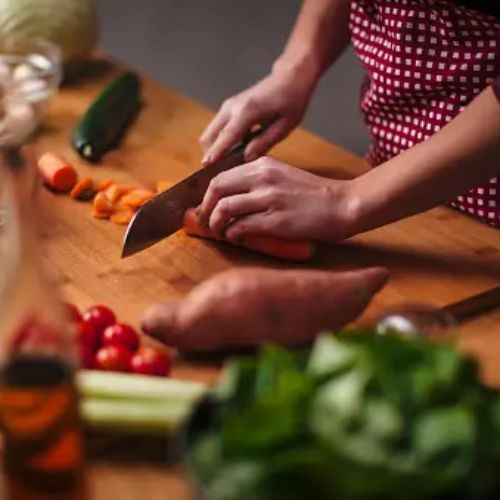 A person chopping vegetables, including carrots and sweet potatoes, on a wooden counter with various fresh ingredients like tomatoes, spinach, and cucumbers laid out nearby.