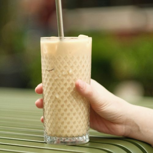 A hand holding a tall glass of iced coffee with a metal straw, resting on a green table with blurred greenery in the background.