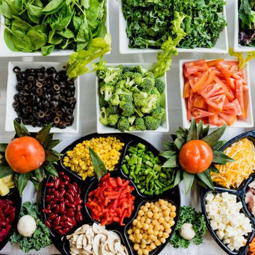 An overhead view of a vibrant salad bar with various fresh vegetables and toppings, including tomatoes, cucumbers, olives, broccoli, chickpeas, corn, and leafy greens arranged in bowls and plates.