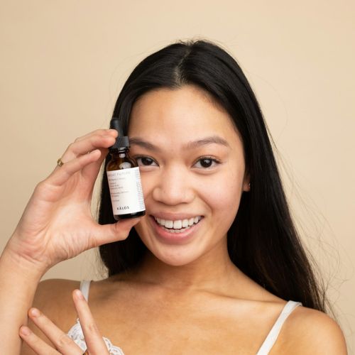 A woman smiling and holding up a small amber glass dropper bottle of facial serum next to their face against a beige background.