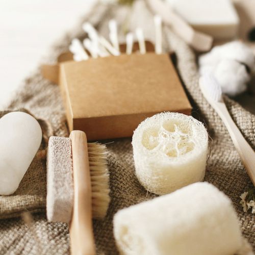 An assortment of natural bath and body items including a loofah sponge, wooden brush, pumice stone, a wooden toothbrush, cotton balls, and a box of cotton swabs arranged on a textured cloth.