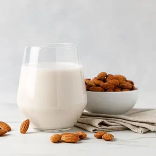 A glass of almond milk is placed next to a bowl filled with almonds on a light-colored napkin. There are a few scattered almonds around the glass and bowl.