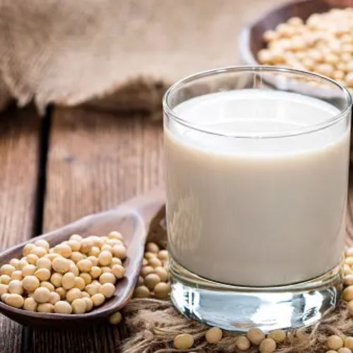 A close-up of a glass of soy milk on a wooden table, accompanied by a wooden scoop and bowls filled with soybeans.