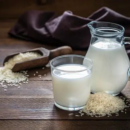 A jug and a glass of rice milk on a wooden table, surrounded by scattered rice grains and a scoop filled with rice. There is a dark cloth in the background.