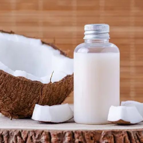 A bottle of coconut milk is set next to a half coconut and coconut pieces on a wooden surface against a background of bamboo-looking material.