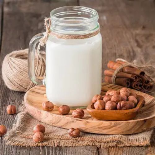 A glass mug filled with hazelnut milk is set on a wooden tray, accompanied by a small bowl of hazelnuts and tied with a piece of twine. Some hazelnuts and cinnamon sticks are scattered around.