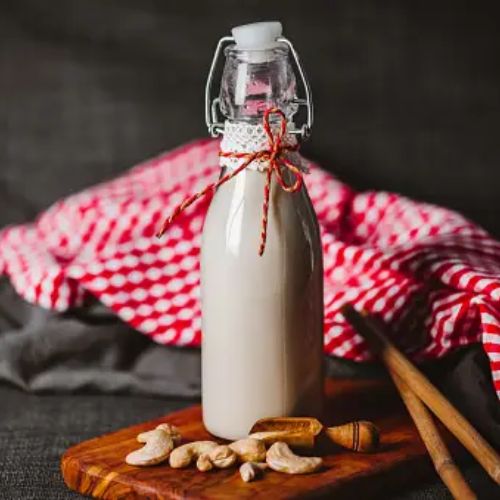 A glass bottle of homemade cashew milk is placed on a wooden board, surrounded by cashews, small wooden utensils, and a red and white checkered cloth in the background.