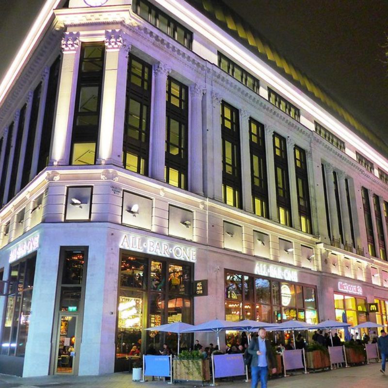 The image shows a corner view of an All Bar One location, with a grand, multi-story building illuminated at night. The outdoor seating area is lively, with patrons sitting under umbrellas, and the building’s elegant architecture is highlighted by exterior lighting.