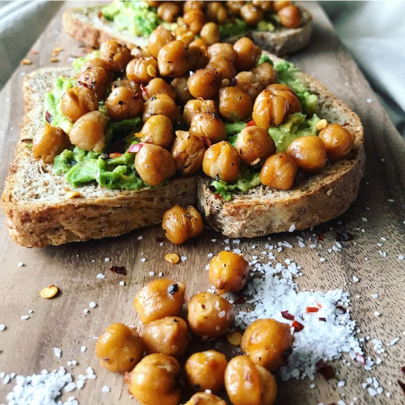 Whole grain toast spread with mashed avocado, topped with roasted chickpeas and sprinkled with red pepper flakes. The toast is served on a wooden board, accompanied by a scattering of coarse sea salt and extra chickpeas.