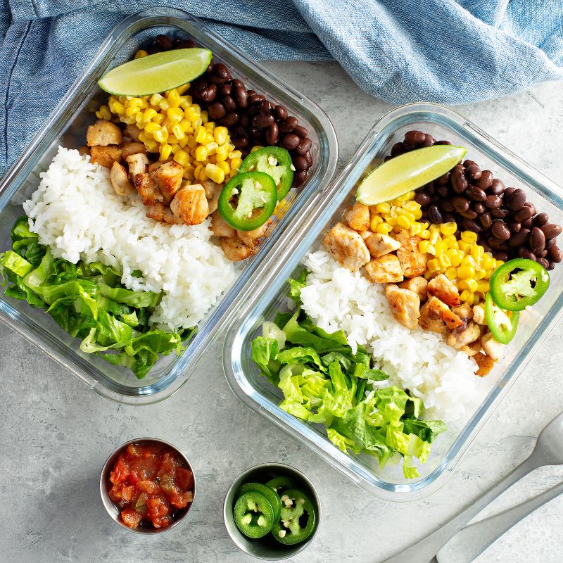Two meal prep containers filled with colourful ingredients, including rice, grilled vegan chicken, black beans, corn, lettuce, jalapeño slices, and lime wedges, accompanied by small bowls of salsa and jalapeños.
