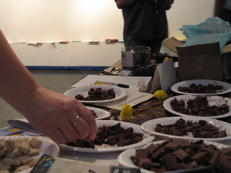 This image shows a table laid out with several plates filled with small squares of chocolate, possibly for a tasting event. A person's hand reaches toward one of the plates, selecting a piece of chocolate. In the background, there are papers, a box, and a jar on the table, along with small decorative yellow chicks. The overall setting suggests a casual, possibly community-based chocolate tasting or event.