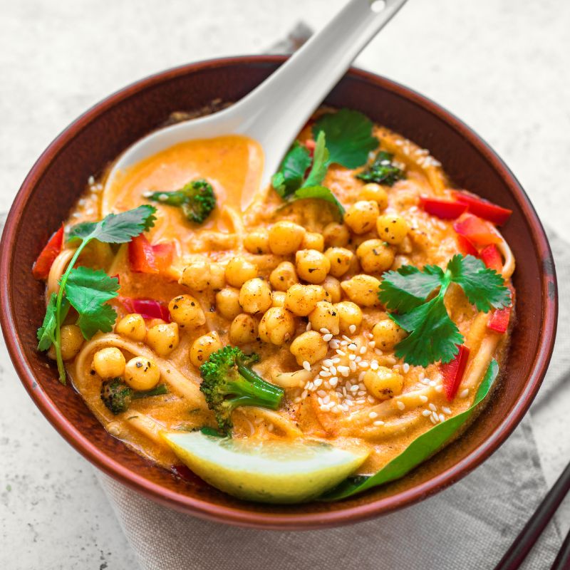 A bowl of chickpea laksa soup, a rich and creamy dish with a spicy, coconut-based broth. The soup is garnished with roasted chickpeas, broccoli, sliced red peppers, fresh cilantro, and a wedge of lime. The vibrant colours and variety of textures make the dish look both hearty and flavourful, with a blend of creamy, crunchy, and fresh elements. A white soup spoon rests in the bowl, ready for serving.