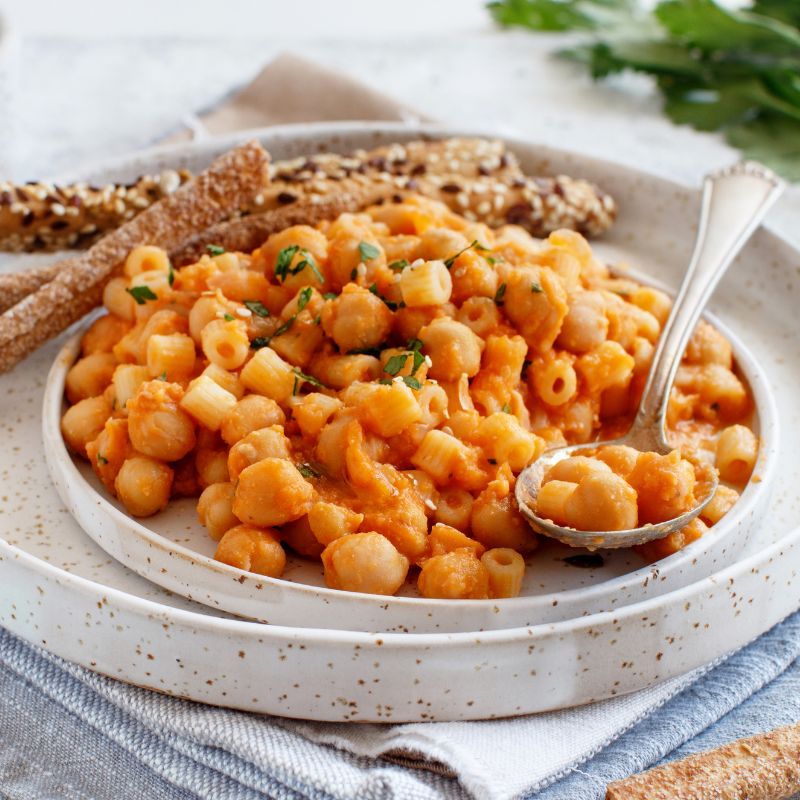 A plate of chickpea pasta in a creamy, orange-coloured sauce, garnished with fresh herbs. The dish features small pasta shapes mixed with whole chickpeas, giving it a hearty and comforting appearance. A spoon rests in the pasta, ready to serve, and the plate is accompanied by seeded breadsticks, adding a crunchy texture to the meal.