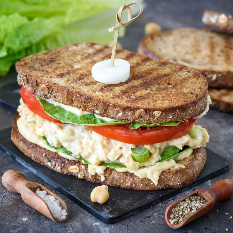 A grilled chickpea salad sandwich made with whole grain bread. The sandwich is layered with a creamy chickpea filling, fresh lettuce, and thick slices of tomato. The bread is toasted, displaying grill marks, and the sandwich is secured with a skewer topped with a slice of pickled onion. The sandwich is served on a dark slate board, with a sprinkle of salt and pepper on the side.