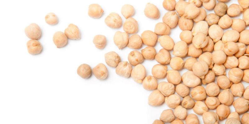 A scattering of dried chickpeas, or garbanzo beans, against a white background. The chickpeas are light beige in colour and have a round, slightly wrinkled texture typical of dried legumes. Some chickpeas are grouped together, while others are spread out, creating a simple and clean presentation.