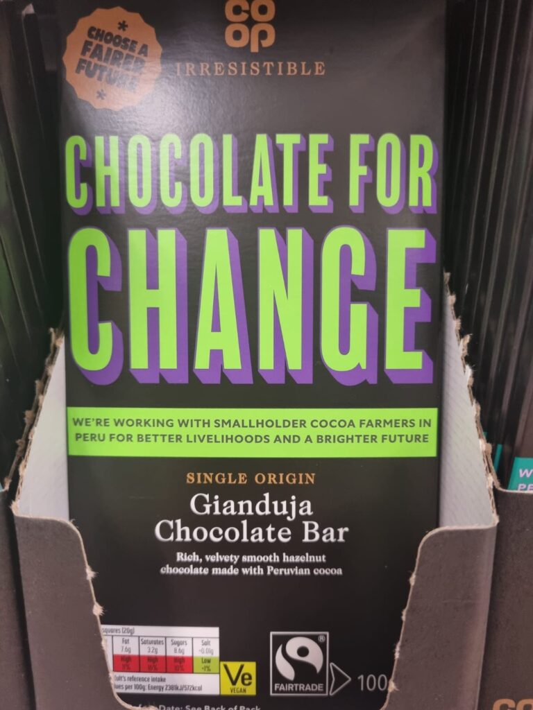 This image shows a Co-op "Chocolate for Change" Gianduja Chocolate Bar on display. The packaging promotes a social message, stating that the company is working with smallholder cocoa farmers in Peru for better livelihoods and a brighter future. The bar is labelled as single-origin, featuring rich and velvety smooth hazelnut chocolate made with Peruvian cocoa. It is Fairtrade certified and vegan, with nutritional information visible at the bottom.