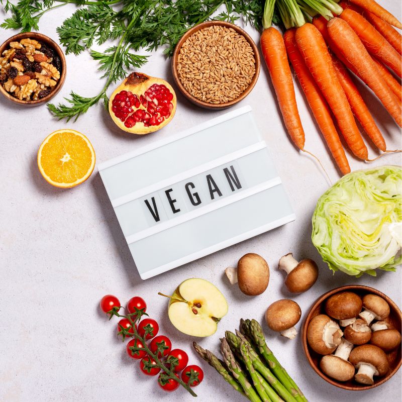A flat lay of fresh produce and ingredients surrounding a sign that says "VEGAN," featuring carrots, lettuce, mushrooms, cherry tomatoes, asparagus, pomegranate, orange slices, walnuts, and grains.