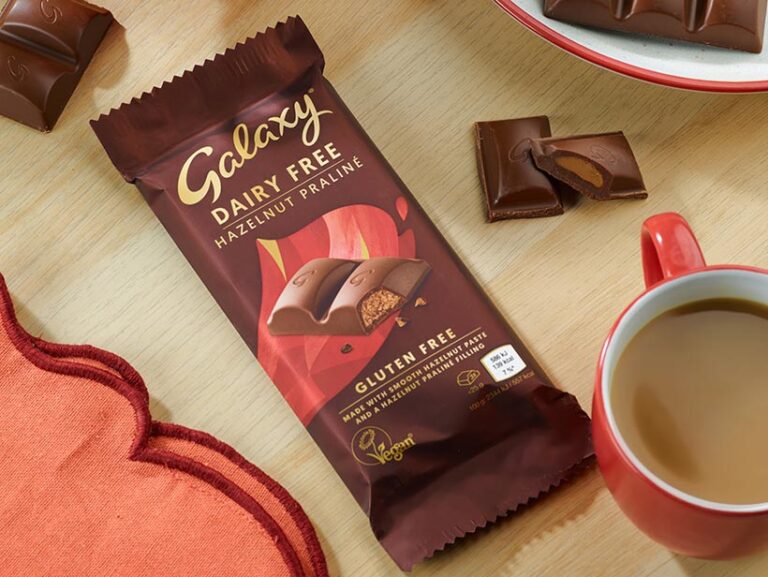 A Galaxy Dairy-Free Hazelnut Praline chocolate bar on a wooden table, next to a red mug filled with coffee and broken chocolate pieces on a plate. The packaging emphasises that it is 'Dairy Free,' 'Gluten Free,' and 'Vegan,' featuring a hazelnut praline filling.