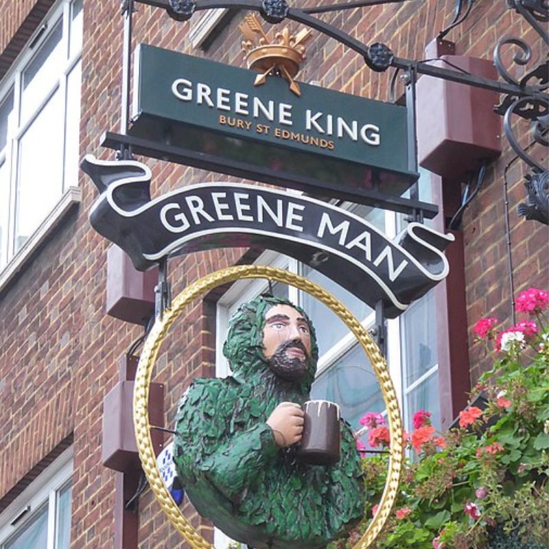This image captures a traditional pub sign for the Greene Man, a part of the Greene King chain, featuring a sculpted figure holding a beer mug. The sign is ornate with floral accents and hangs outside a brick building, adding a classic, British pub feel.