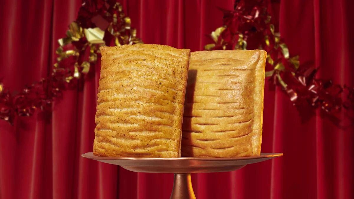 Two Greggs festive vegan bakes on display, set against a festive backdrop with red curtains and gold decorations, highlighting the warm, golden-brown flaky crust of the pastries as they sit atop a pedestal.