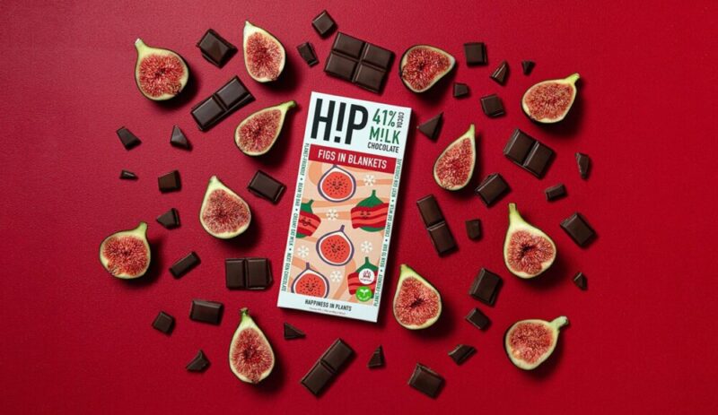A flat lay of a "HiP 41% Milk Chocolate" bar with the flavour "Figs in Blankets" surrounded by halved fresh figs and scattered pieces of dark chocolate on a vibrant red background. The arrangement highlights the bar's plant-based and vegan-friendly branding.